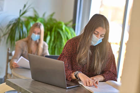 Twins sisters are wearing protective mask and are working together their lessons on their laptop at home during the lockdownの写真素材