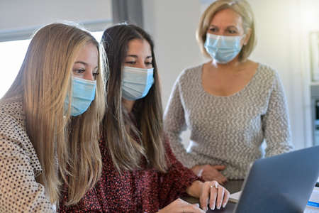 Twin sisters and their teacher wearing protective mask and working their lesson on a laptop at home during lockdown due to covid-19の写真素材