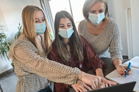 Twin sisters and their mother wearing protective mask and watching a webinar on a laptop at home during lockdown due to covid-19の写真素材