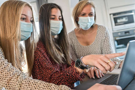 Twin sisters and their teacher wearing protective mask and working their lesson on a laptop at home during lockdown due to covid-19の写真素材
