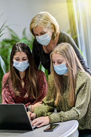 Twin sisters and their mother wearing protective mask and watching a webinar on a laptop at home during lockdown due to covid-19の写真素材