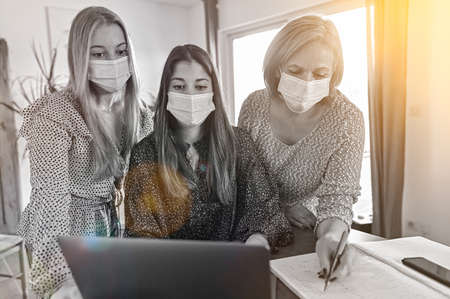 Twin sisters and their mother wearing protective mask and working a elearning lesson on a laptop at home during lockdownの写真素材