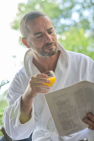 Portrait of a man wearing a bathrobe and reading the morning news on a newspaper while he is drinking his orange juice at the breakfastの写真素材