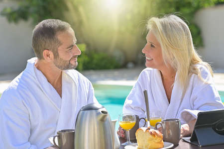 Mature couple having breakfast in bathrobe outside in front of their swimming poolの写真素材