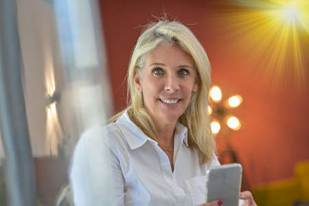 Portrait of a blonde woman in his living room on his mobile phone, surfing the web and reading her messagesの写真素材