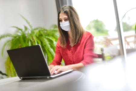 Young female woman wearing a protective mask while she is working at home and attending at a meeting on her computer during the lockdownの写真素材