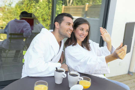 Happy young couple in bathrobes having breakfast outdoors while they are on doing a video callの写真素材