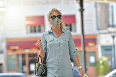Attractive middle age urban woman walking in the street and carrying bags for shopping while she is wearing a protective maskの写真素材