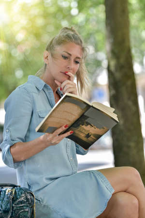Woman doing a break outdoors in a park, setting on a bench and reading a bookの写真素材