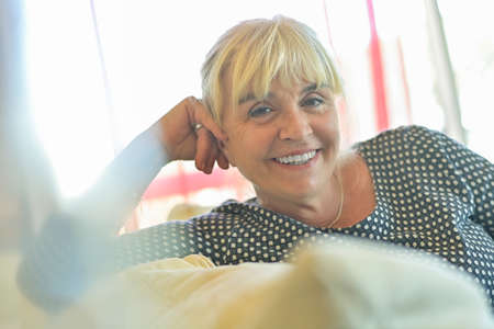 Portrait of a smiling mature blond woman sitting in her sofaの写真素材