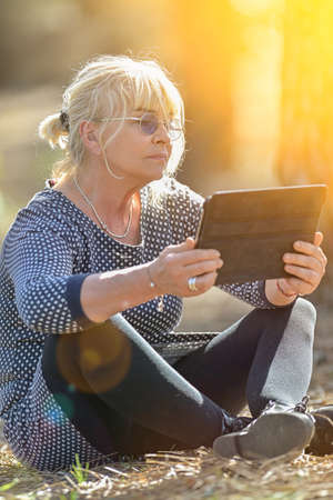 Mature woman doing a break outdoors in a park, setting on the floor and reading a digital bookの写真素材
