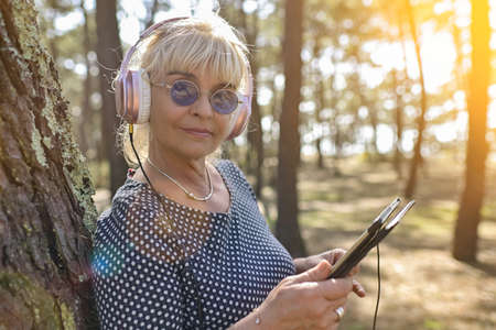Mature blonde woman standing outdoors in a park listening to music on her digital tablet with her headphones.の写真素材
