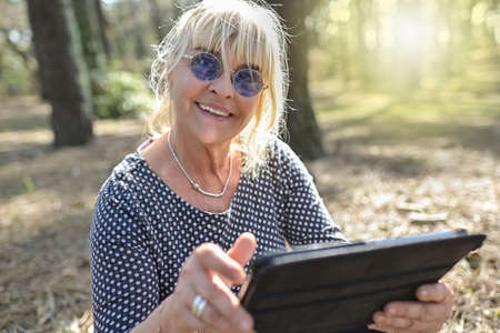 Mature woman doing a break outdoors in a park, setting on the floor and reading a digital bookの写真素材