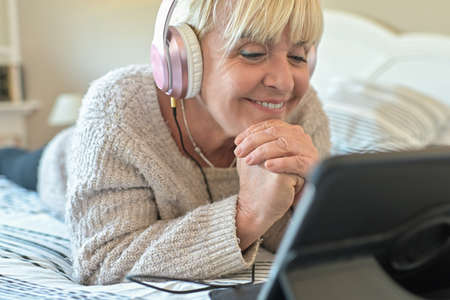 Mature blonde woman lying on her sofa and listening to music on her digital tablet with her headphones.の写真素材