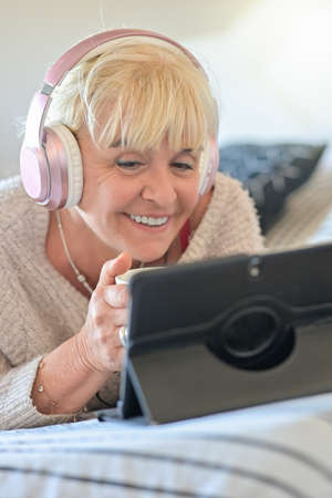 Mature blonde woman lying on her sofa dicussing with her friends on a video call on her digital tablet with her headphones.の写真素材
