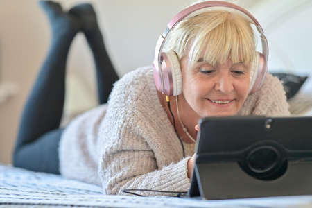 Mature blonde woman lying on her sofa and listening to music on her digital tablet with her headphones.の写真素材