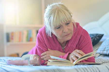 Portrait of a mature retired woman lying on her bed, relaxed and reading her bookの写真素材