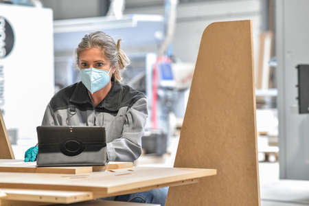Woman reading technical information on her tablet in an industrial carpenter workshop and wearing protections against virusの写真素材