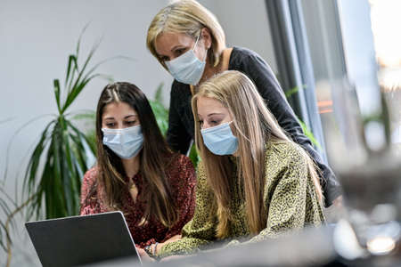 Twin sisters and their mother wearing protective mask and watching a webinar on a laptop at home during lockdown due to pandemicの写真素材
