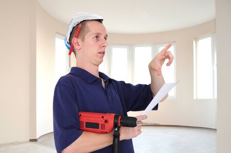 Young serious man with drill, blueprints and paper hat pointing in new appartmentの写真素材