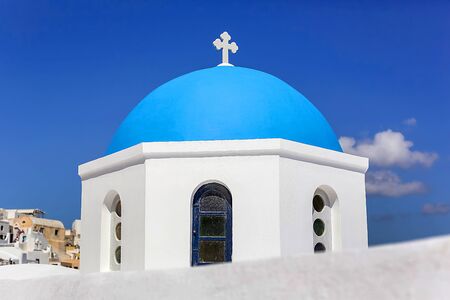 Close-up of the round dome of the Church with a blue cross, white stone walls against the blue sky in Greece on Santoriniの写真素材
