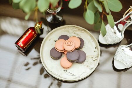 Close-up of the bride's accessories, white shoes open sandals, two gold wedding rings, a bottle of red perfume lying on a black table. A lot of macaroon cakes pink and lilac on a white round plateの写真素材