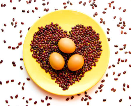 Three brown eggs on red beans shaped as heart on a yellow plateの写真素材