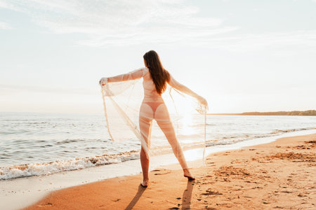 Young woman in red swimsuit and transparent cape walk along sandy beach near seaの写真素材