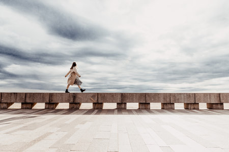 Young woman in autumn coat walking along an empty embankment on rainy cloudy day, cityscapeの写真素材