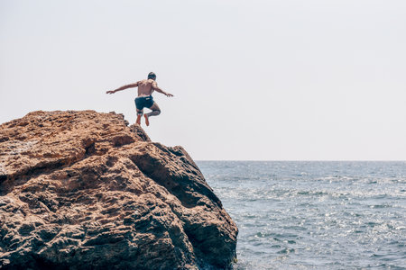 Man in swimsuit jumping from large stone into water sea, landscape on summer dayの写真素材