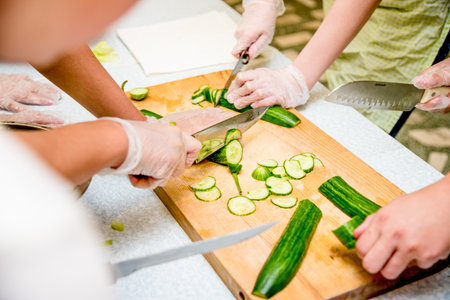 People in kitchen preparing vegetable salad, close-up hands cutting cucumber on cutting boardの写真素材