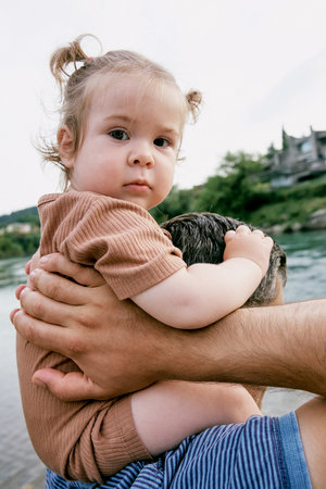 Little cute girl toddler daughter sitting on dad's shoulders, family walking on pebble river beachの写真素材