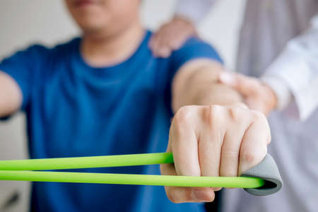 Doctor physiotherapist assisting a male patient while giving exercising treatment on stretching his arm with exercise band in the clinic, Rehabilitation physiotherapy concept.の写真素材