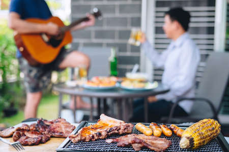 Group of friends Two young man enjoying grilled meat and play guitar with raise a glass of beer to celebrate the holiday festival happy drinking beer outdoors and enjoyment at home.の写真素材
