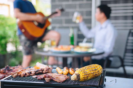 Group of friends Two young man enjoying grilled meat and play guitar with raise a glass of beer to celebrate the holiday festival happy drinking beer outdoors and enjoyment at home.の写真素材