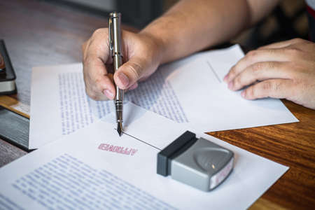Close up hands of businessman signing and stamp on paper document to approve business investment contract agreement.の写真素材