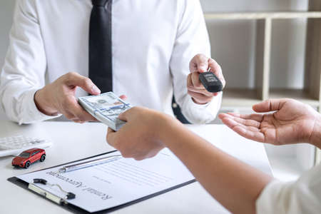 Realtor agent manager holding key of new car giving to woman client after signing rental contract form and sending dollar us banknotes, renting considering vehicle car insurance.の写真素材
