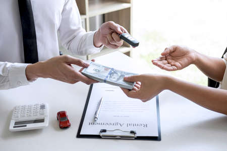 Realtor agent manager holding key of new car giving to woman client after signing rental contract form and sending dollar us banknotes, renting considering vehicle car insurance.の写真素材