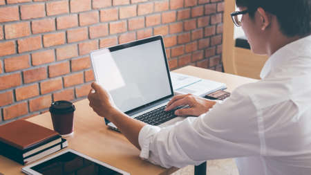 Image of Young man working in front of the laptop looking at screen with a clean white screen and blank space for text and hand typing information on keyboard in modern workspace.の写真素材