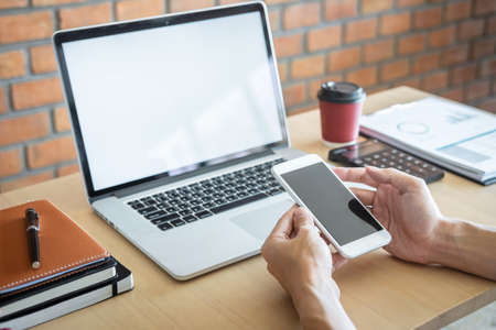 Image of Young man working in front of the laptop looking at screen with a clean white screen and blank space for text, hand holding smartphone and typing information on keyboard in modern workspace.の写真素材