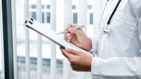 The doctor is standing and reading medical report inside his office, the doctor is reading the patient's symptoms and analyze treatment, Health care and medicine used to treat the patientの写真素材