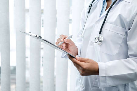 The doctor is standing and reading medical report inside his office, the doctor is reading the patient's symptoms and analyze treatment, Healthcare and medicine used to treat the patientの写真素材