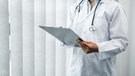 The doctor is standing and reading medical report inside his office, the doctor is reading the patient's symptoms and analyze treatment, Health care and medicine used to treat the patientの写真素材