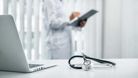 Doctor is standing and reading report and Stethoscope is on the table, doctor is reading symptoms of the patient and analyze treatment, health care and medicine at his hospital officeの写真素材