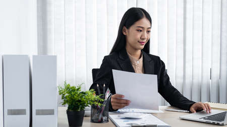Young secretary woman holding paper document to typing email to her boss and clients while sitting...