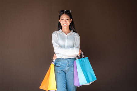 Portrait of asian shopaholic woman smiling and wearing sunglasses headband with many colorful shopping bags in both hands after heavy shopping isolated over brown backgroundの写真素材