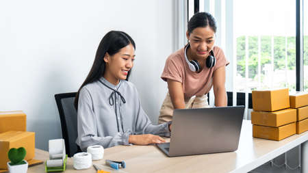 Two young business woman entrepreneur are checking order together on laptop and smiling while talking with client using smartphone, couriers are preparing packing product to parcel box home deliveryの写真素材