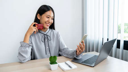 Young asian woman smiling and showing credit card in her hands for online shopping while looking on smartphone to using mobile banking and entering security authorization codeの写真素材