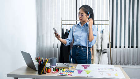 Asian woman fashion designer looking video on smartphone and thinking new clothes collection while standing to working at her workshopの写真素材