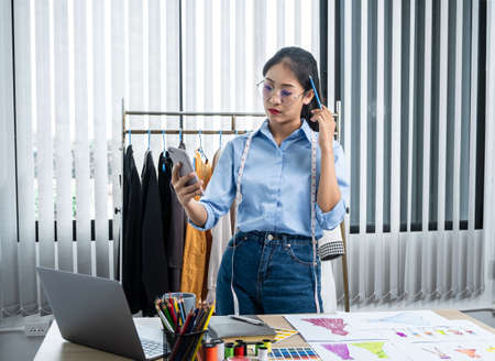 Asian woman fashion designer looking video on smartphone and thinking new clothes collection while standing to working at her workshopの写真素材